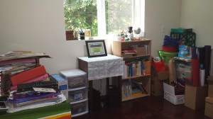 A storage unit I bought after the fire, the old sewing machine my friends gave me, a cheap bookcase Tom acquired while we lived in the apartment.  The striped cloth protecting the top of the desk was at school when I had the fire, and references the stripes on the bookcase.  The colors make me happy.  The collection of boxes and bags on the right-hand wall makes me cringe...