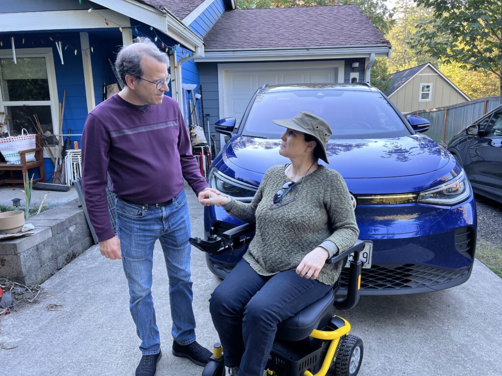 two people holding hands, one standing one in wheelchair, in front of a car in the driveway