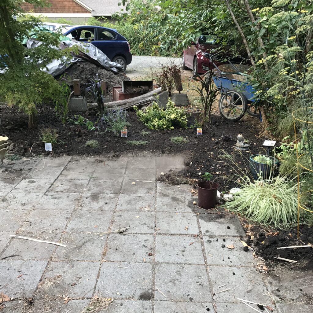 image of the enlarged patio in front with staggered edges going up to the planting beds. A large pile of soil under a top is in the background.